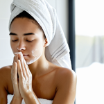 A young adult woman, fully clothed in a modest, soft, white towel wrap, gently washing her face with natural soap in a brightly lit, minimalist bathroom. Her eyes are closed in a serene expression, a soft, fine foam is visible on her face. Her skin appears hydrated and calm. Natural light streams from a window. Perfect anatomy, correct proportions, natural pose, well-formed hands, proper finger count, natural body proportions. Professional studio photography, high quality, soft lighting, focus on healthy skin, safe for work, appropriate content, fully clothed, modest, family-friendly.